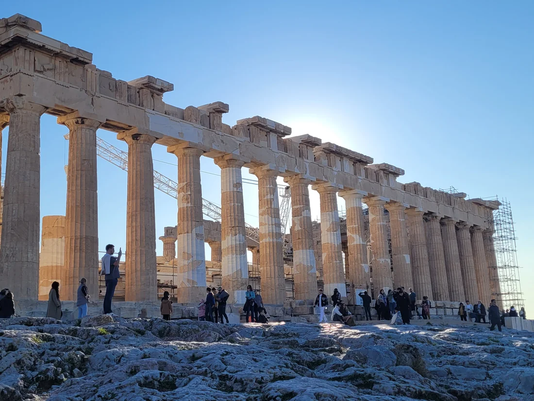 The Parthenon in Athens, backlit by the sun, with visitors on the rocks below