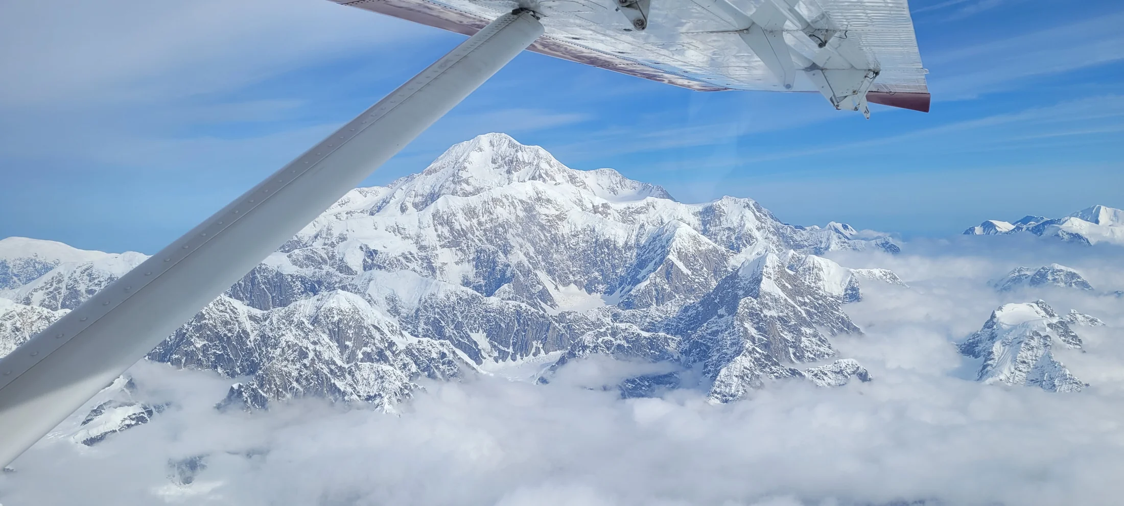 A small plane's wing over Denali's snow-capped peak, above the clouds